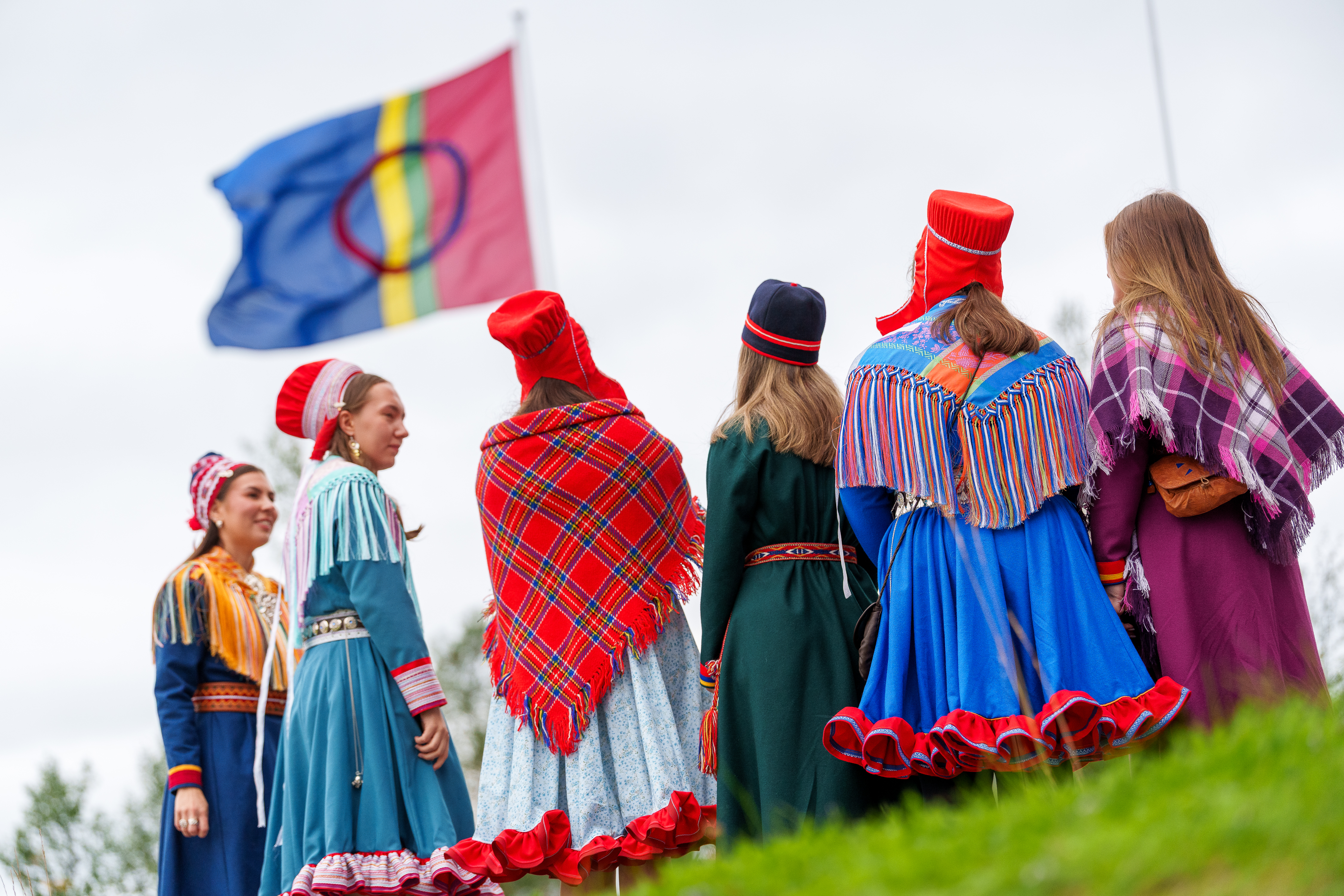 Koftekledde studenter står med ryggen vendt mot kamera. Samisk flagg i bakgrunnen.